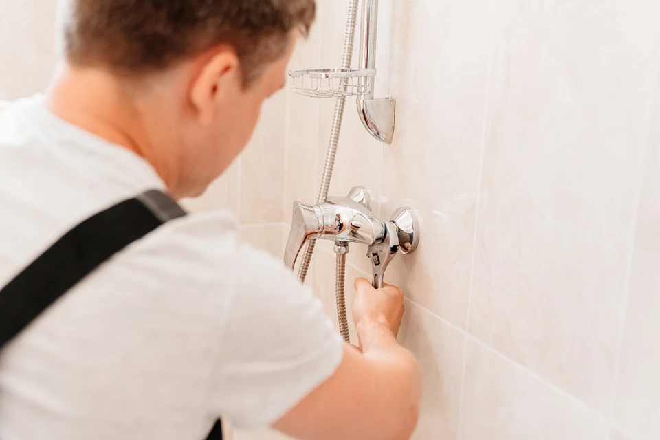 Un plombier en salopette installe ou répare une robinetterie de douche dans une salle de bain carrelée blanche