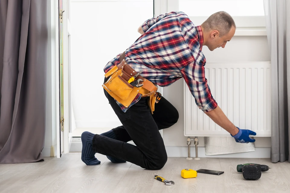 Un plombier ou technicien de chauffage intervient à genoux devant un radiateur blanc dans un intérieur résidentiel
