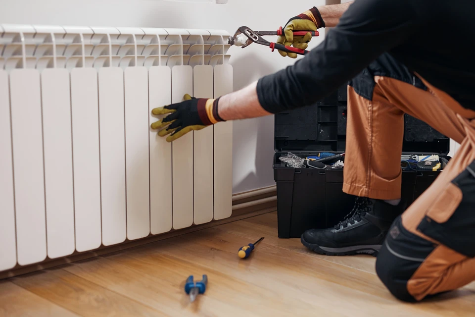Homme installant un radiateur à panneaux dans une maison en rénovation, agenouillé sur parquet bois, murs en plâtre avec briques apparentes