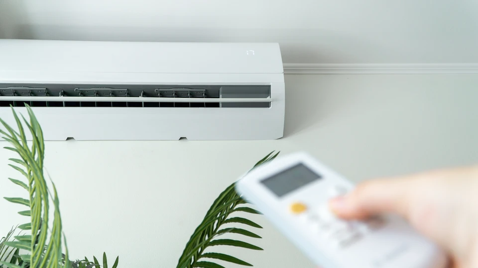 Une main tient une télécommande blanche de climatisation avec écran LCD et boutons de contrôle, dirigée vers un climatiseur mural en arrière-plan dans un intérieur moderne aux murs blancs