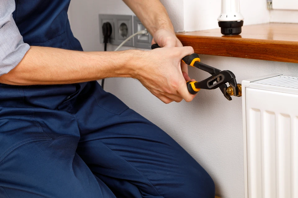 Un plombier ou technicien de chauffage intervient à genoux devant un radiateur blanc dans un intérieur résidentiel
